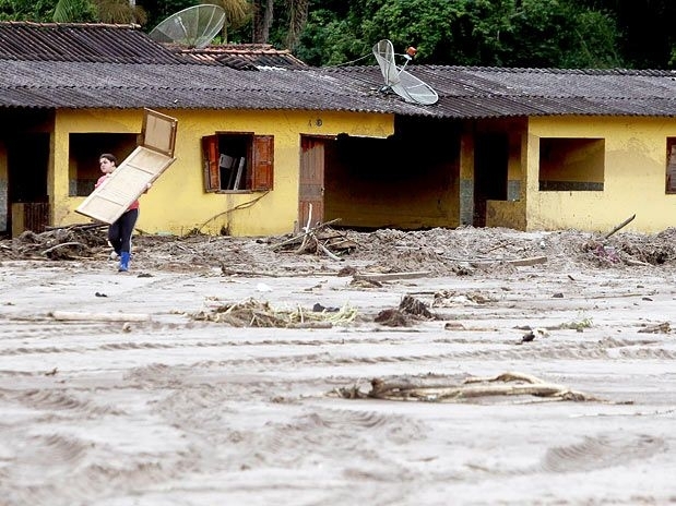 Sumidouro/RJ: mulher carrega porta enquanto atravessa lamaal que restou em frente a casas danificadas por deslizamentos